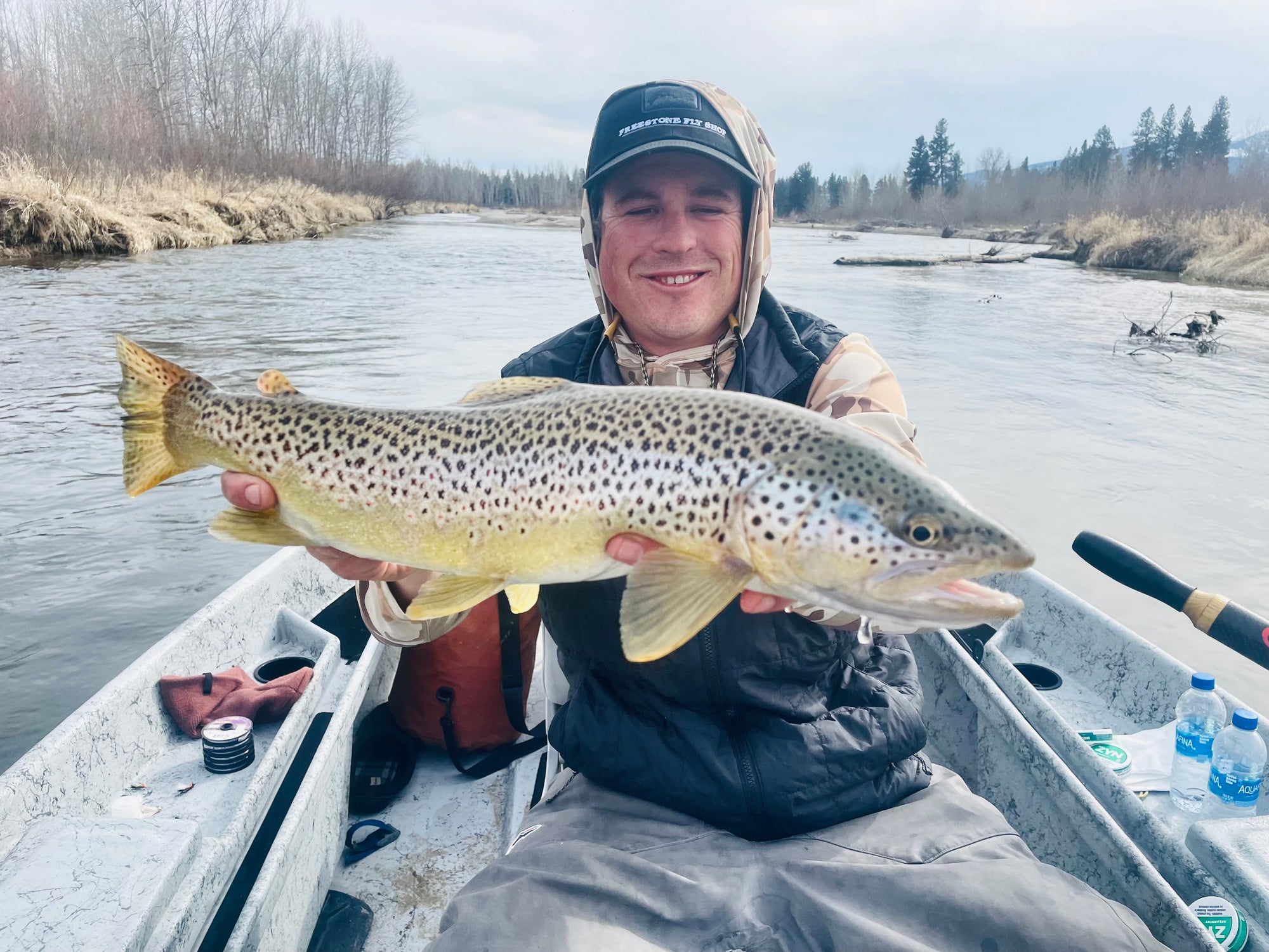 Angler holding large brown trout sitting in small fishing boat on calm river with trees and overcast sky